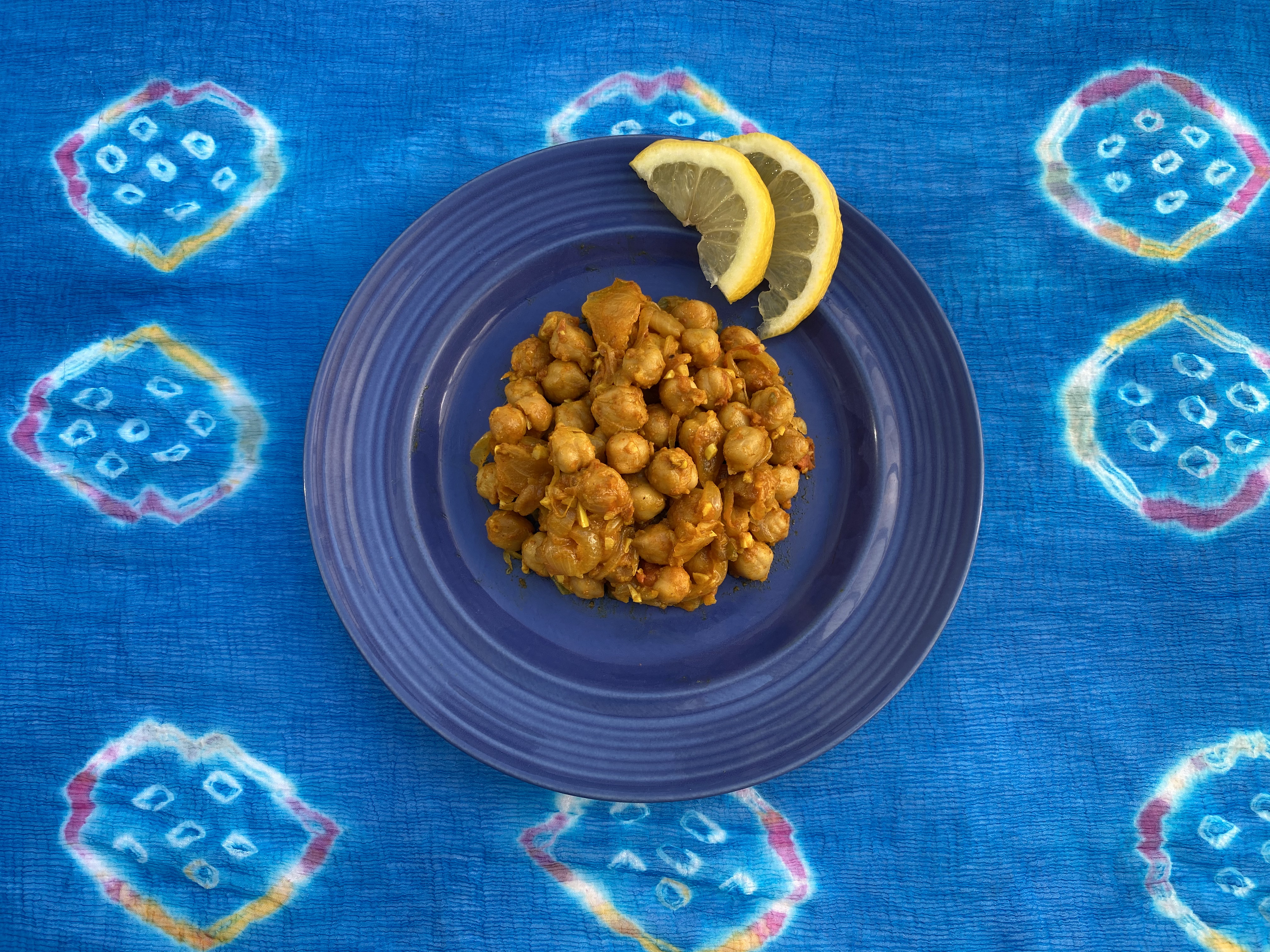 Chickpea Curry served on a blue plate and garnished with lemon slices, against a multi-colored tablecloth.