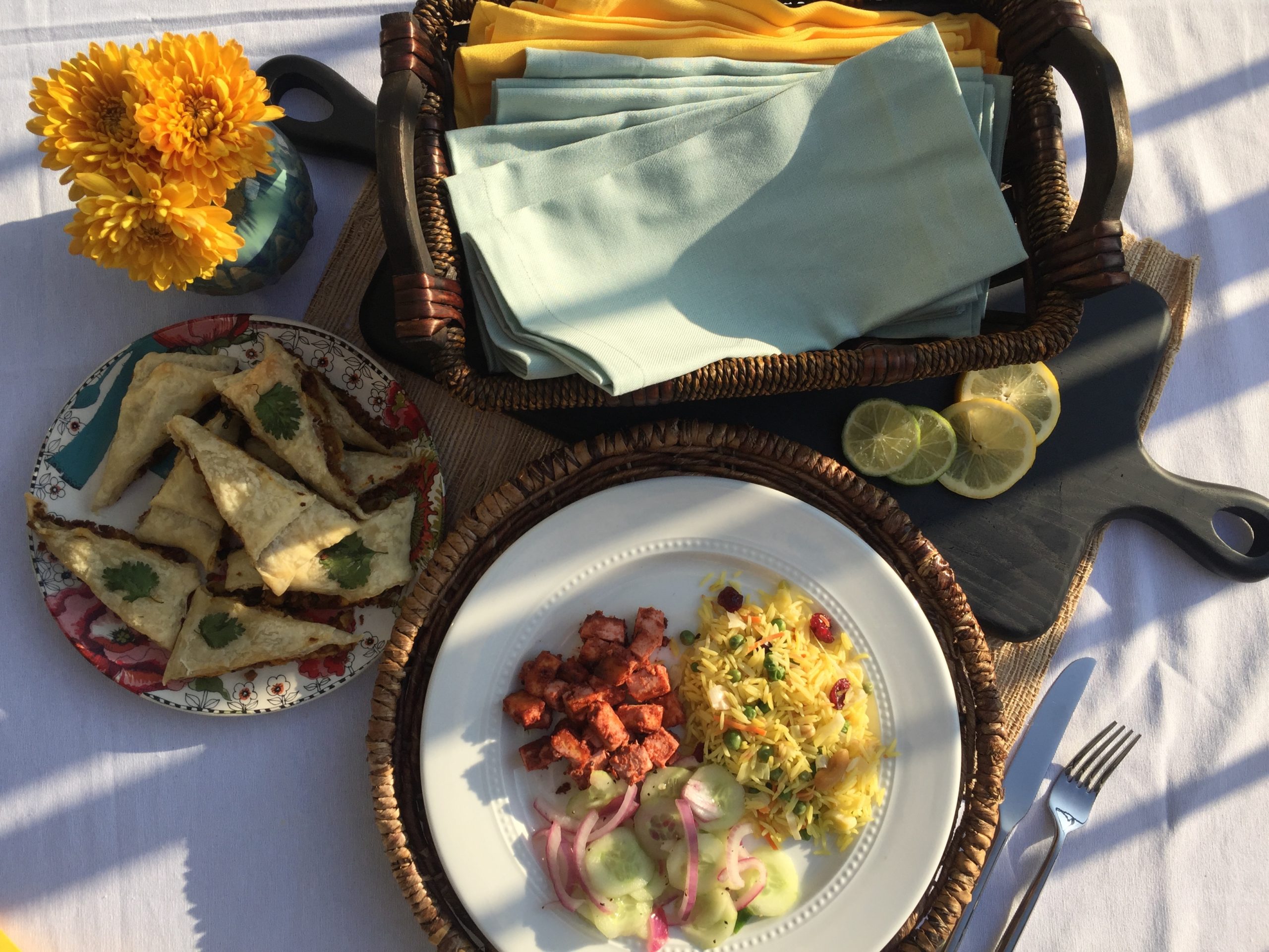 Tandoori Tofu, Yellow Rice Pilaf and Cucumber Salad served on a white plate. On another multi-colored plate are baked samosas. Yellow flowers, basket of napkins and black cutting board with lemon and lime slices as decorative elements.