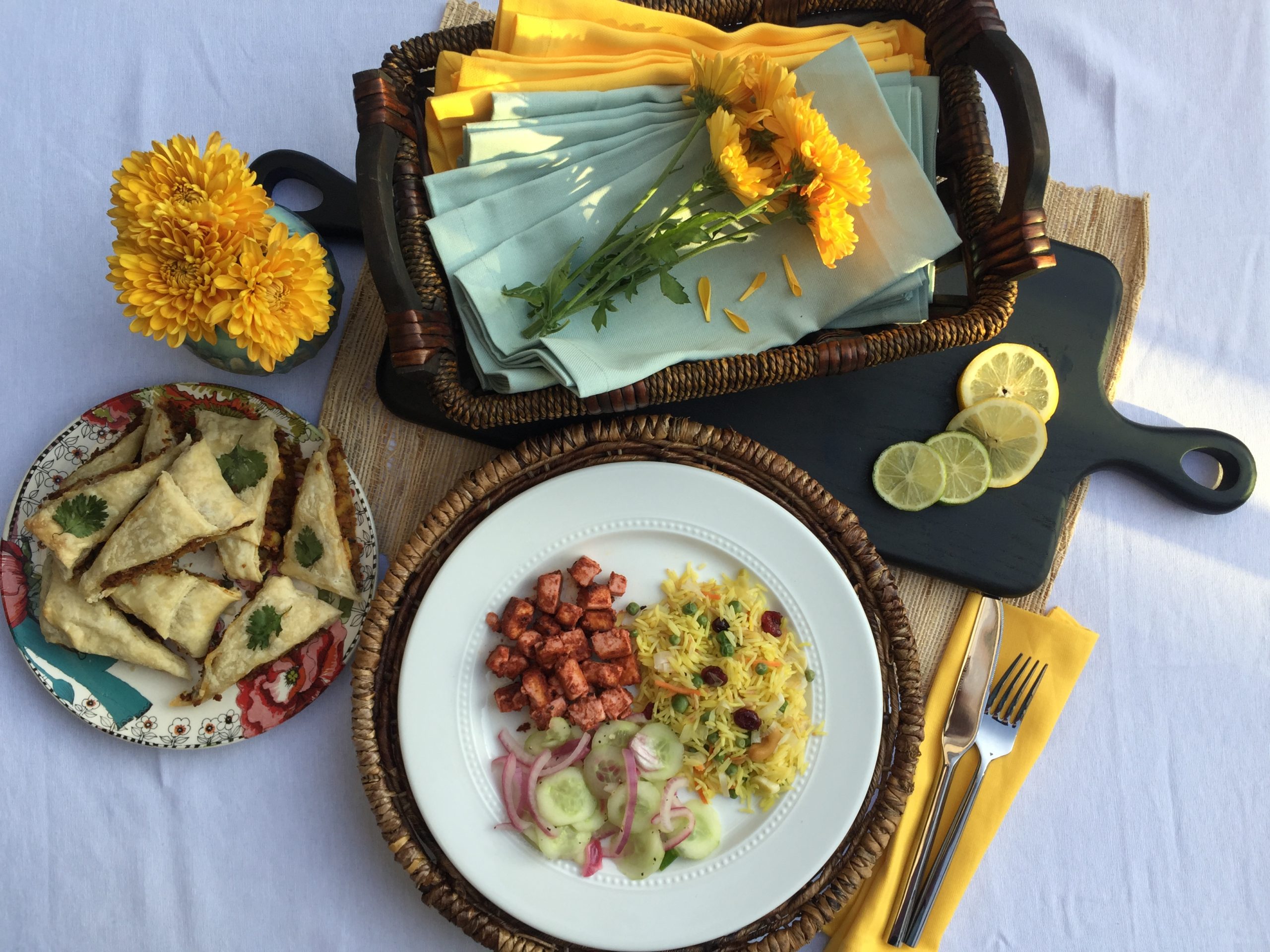 Tandoori Tofu, Yellow Rice Pilaf and Cucumber Salad served on a white plate. On another multi-colored plate are baked samosas. Yellow flowers, basket of napkins and black cutting board with lemon and lime slices as decorative elements.