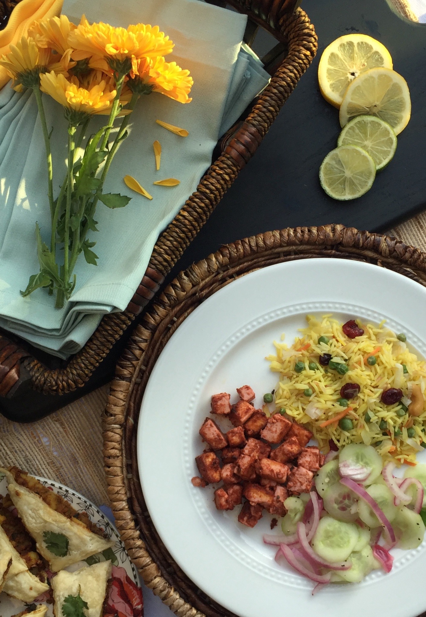 Tandoori Tofu, Yellow Rice Pilaf and Cucumber Salad served on a white plate. On another multi-colored plate are baked samosas. Yellow flowers, basket of napkins and black cutting board with lemon and lime slices as decorative elements.