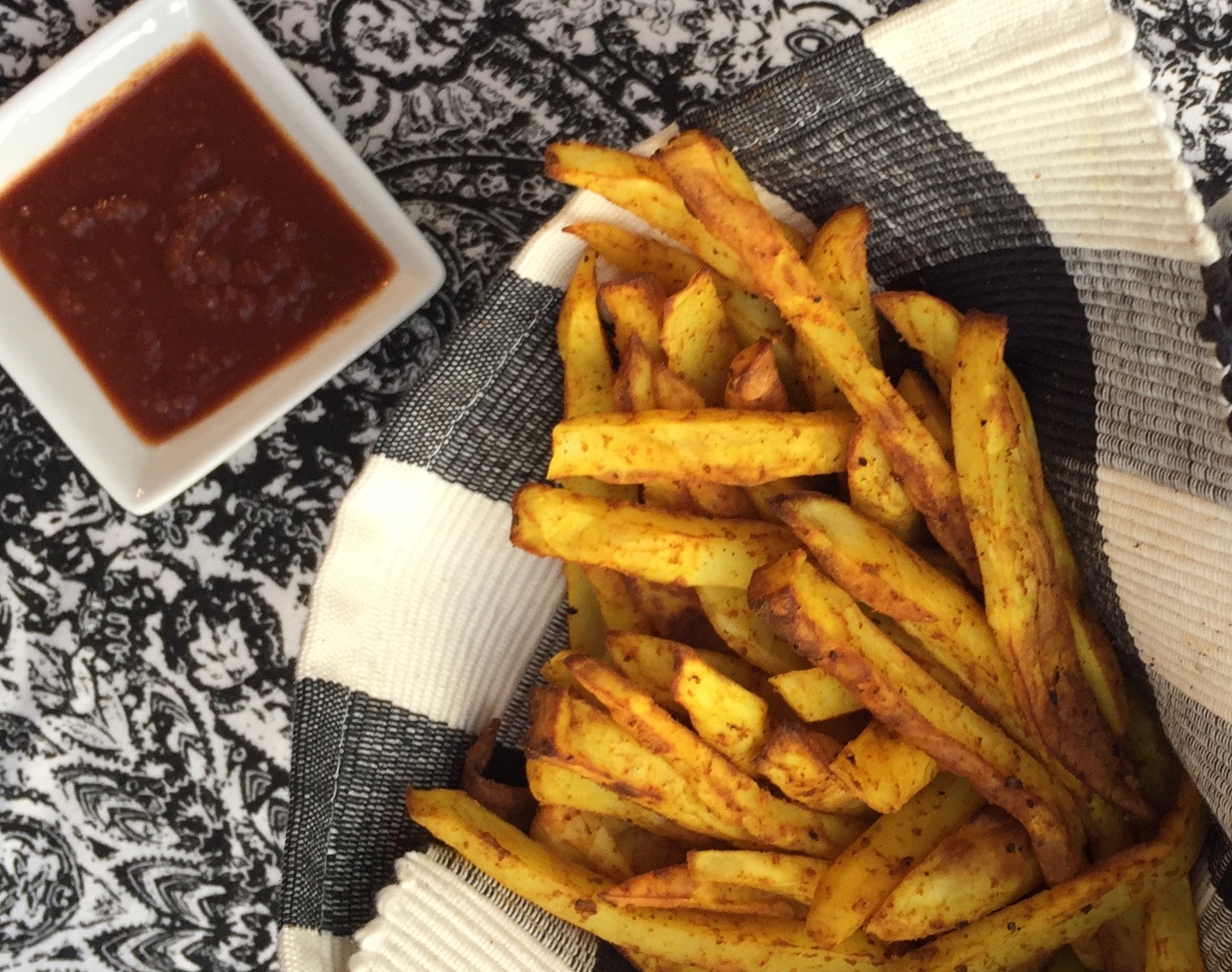 Baked French fries in a black-and-white napkin. A black-and-white tablecloth and a small dish of ketchup are in the background..