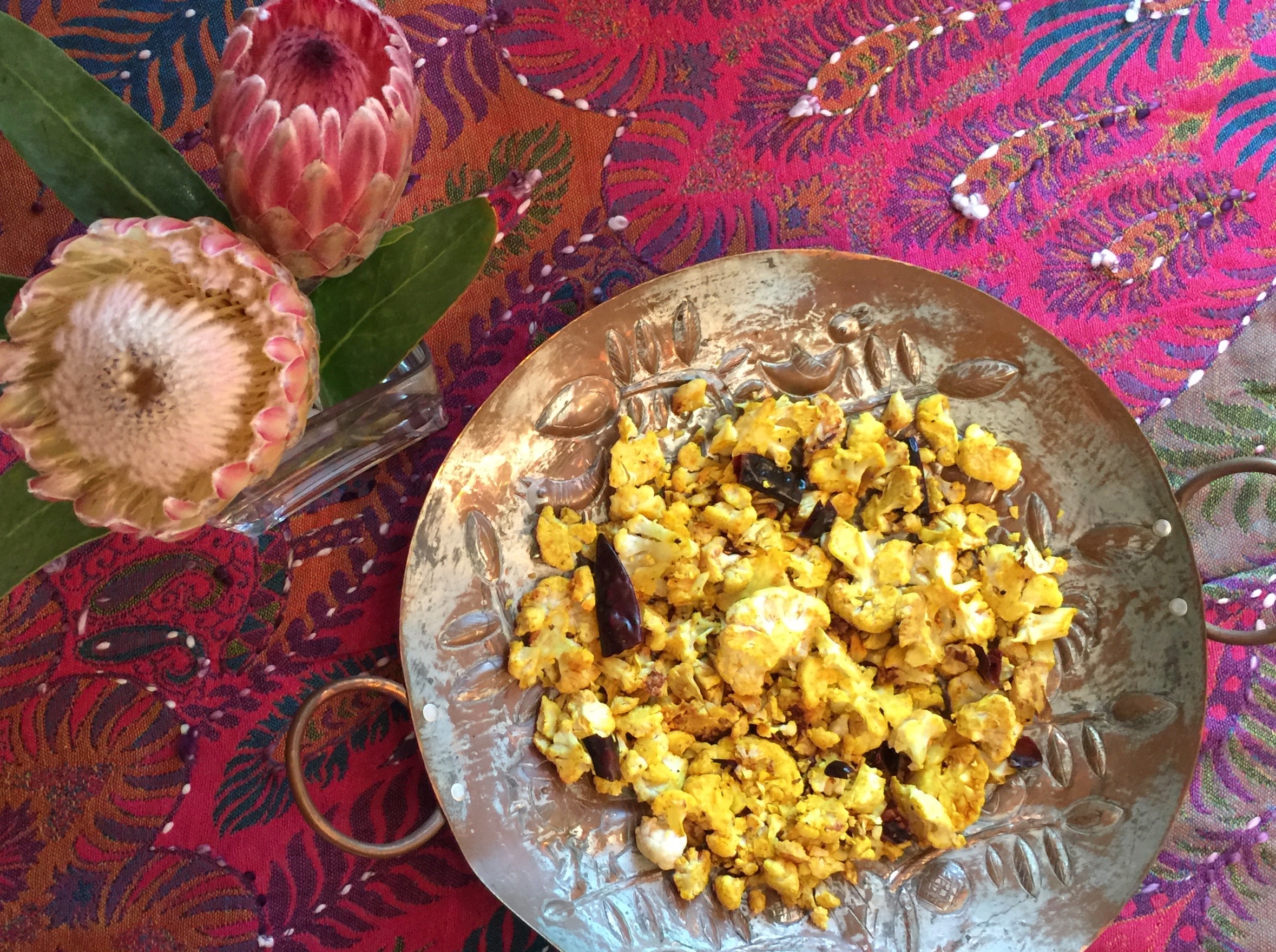 Pan-roasted cauliflower in a silver serving dish. Multi-colored tablecloth and a vase of pink and white flowers