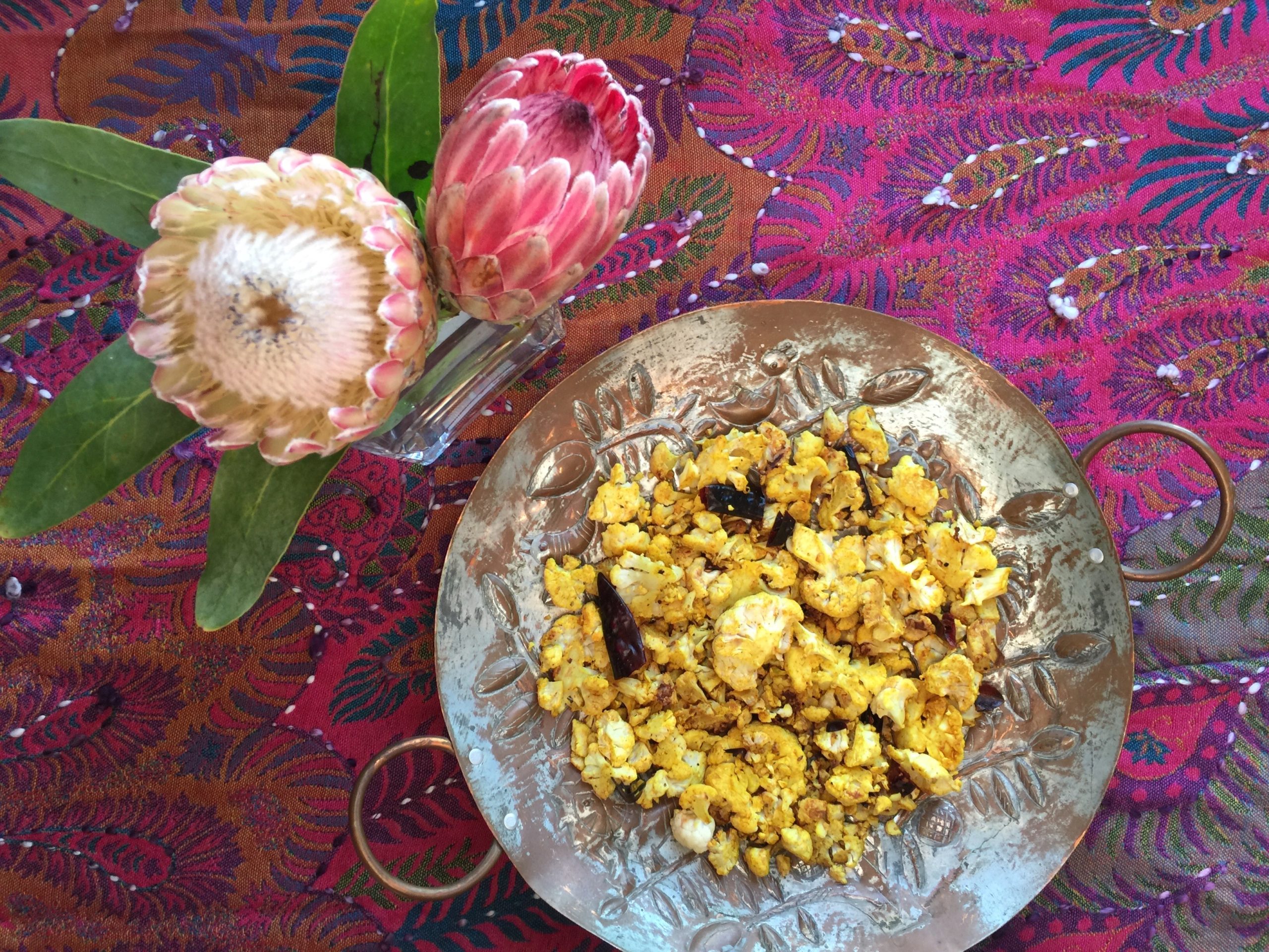 Pan-roasted cauliflower in a silver serving dish. Multi-colored tablecloth and a vase of pink and white flowers