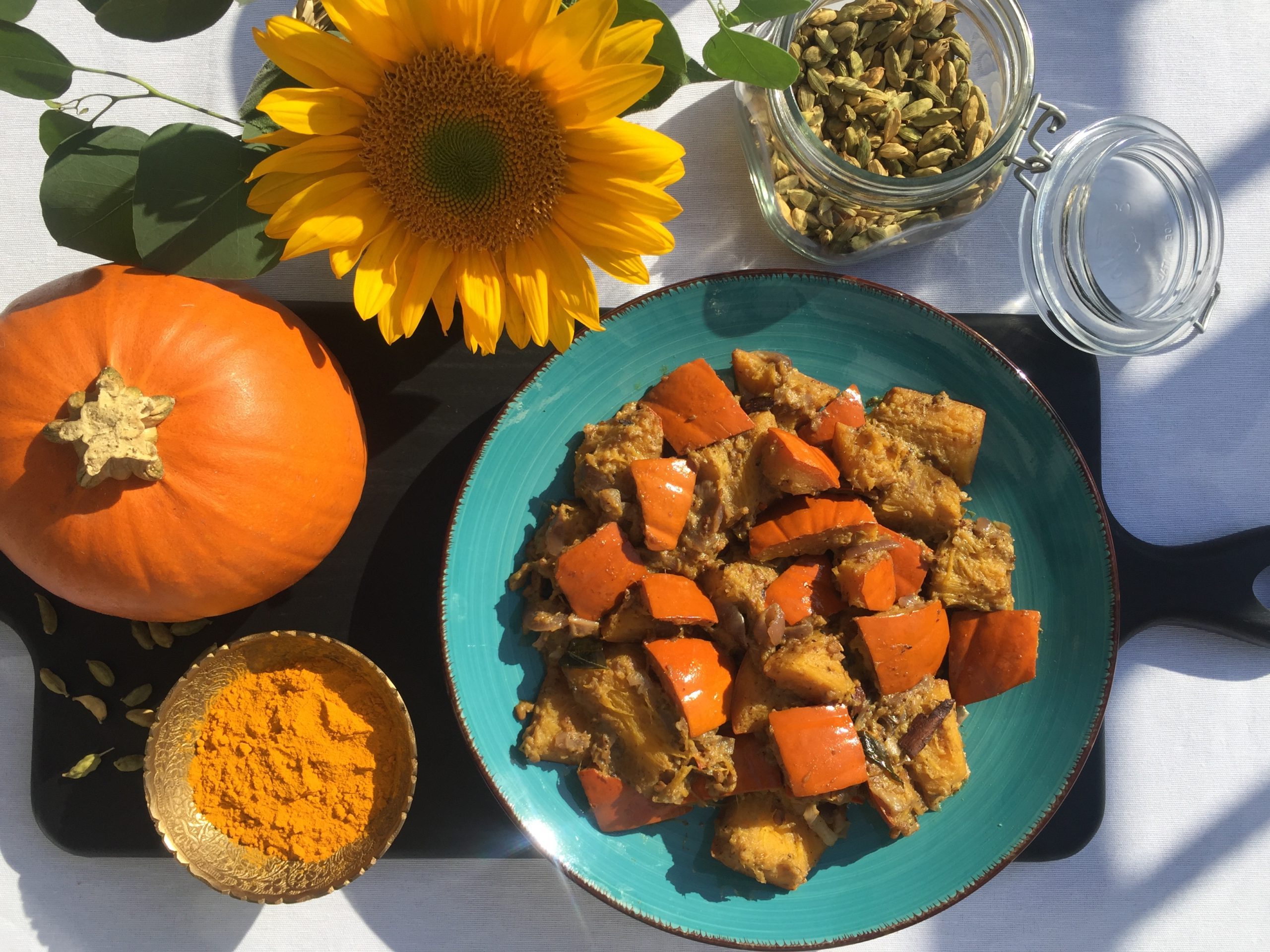 Sri Lankan Pumpkin Curry served on a turquoise plate. Cardamom pods, sunflower, pumpkin and small dish of turmeric as decorative elements.