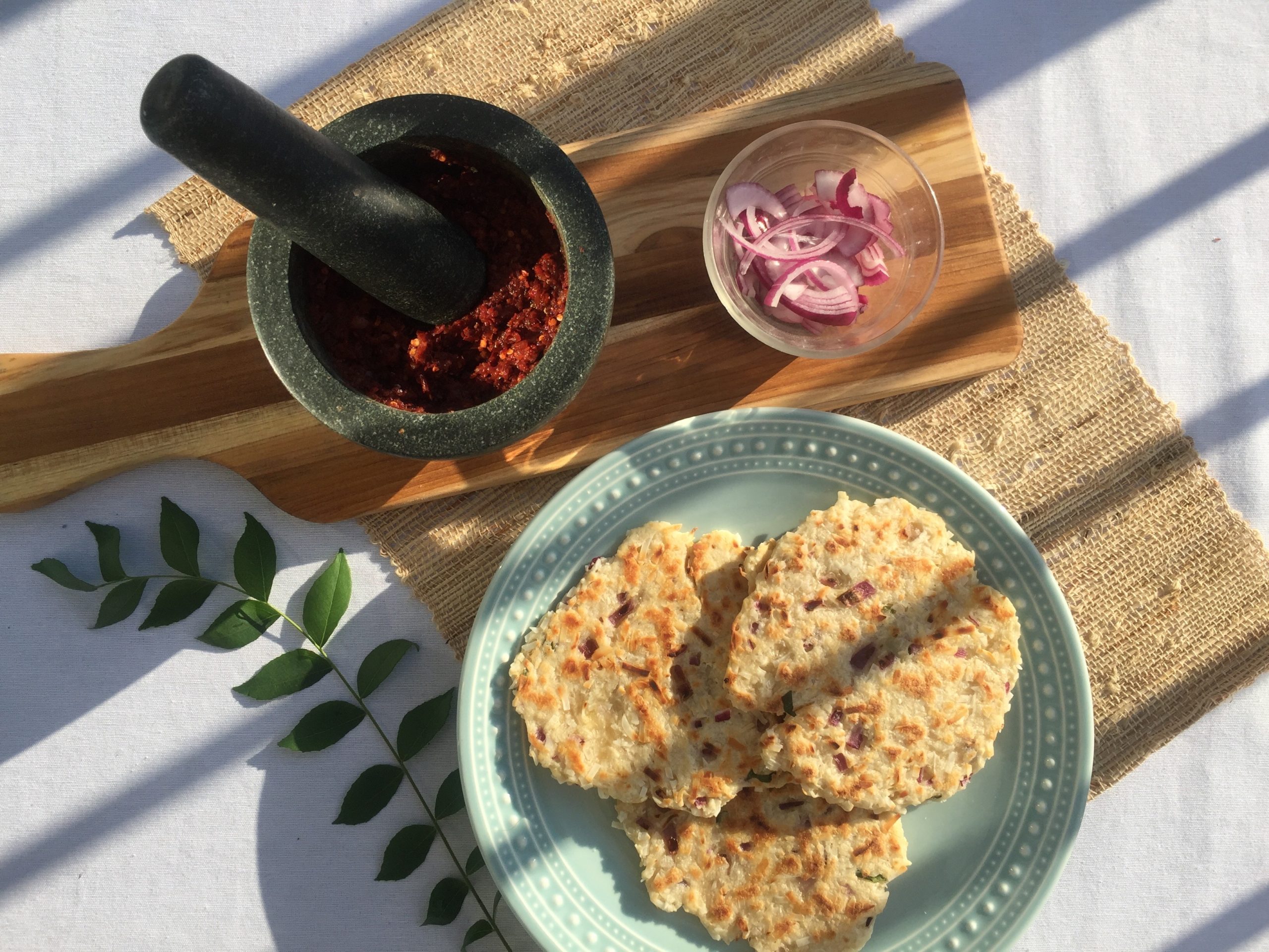 Three roti served on a plate. Mortar containing chili and onion sambol and the pestle. Small dish of sliced onion and sprig of curry leaf as decorative elements.