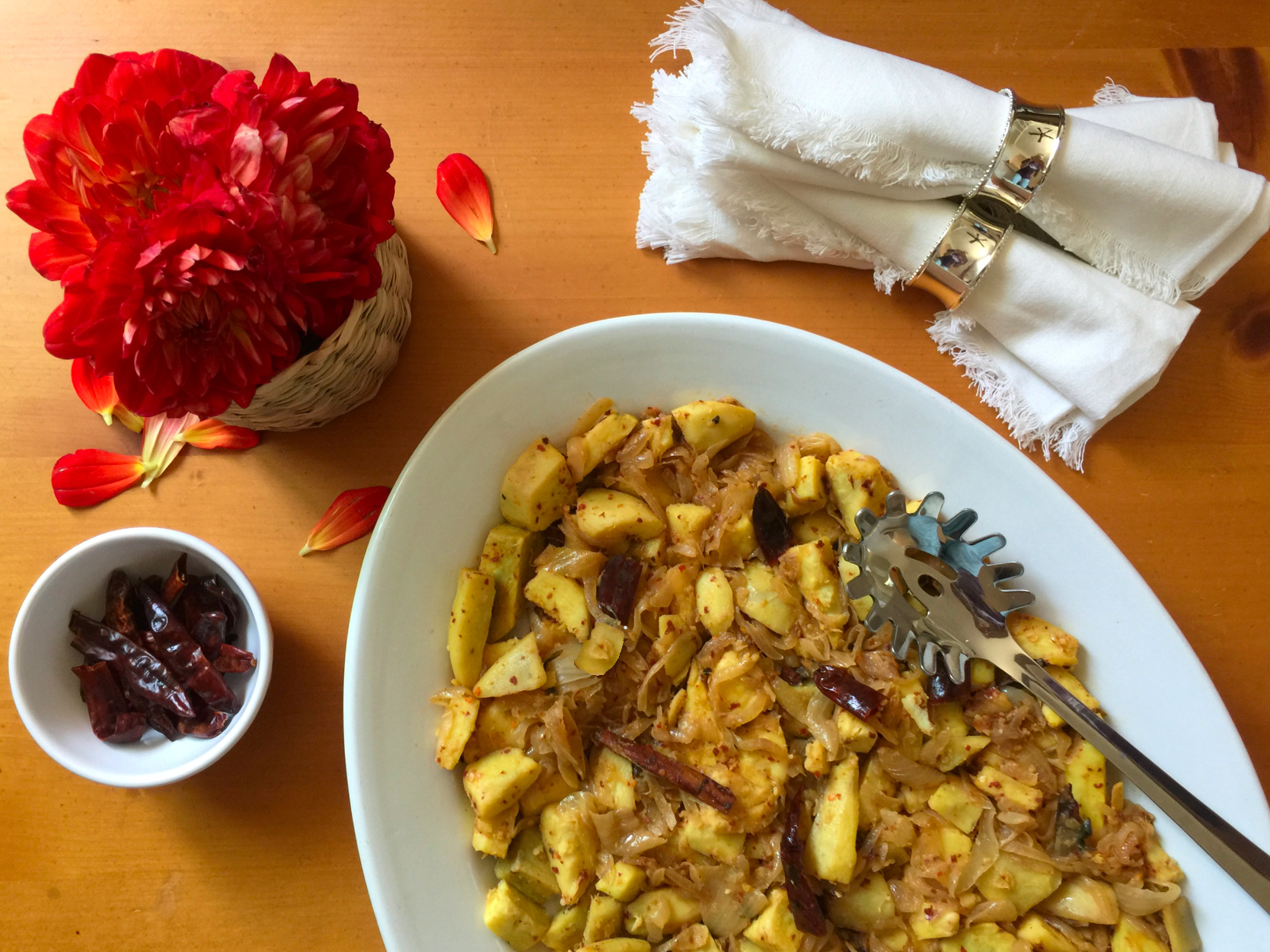 Murasaki Sweet Potatoes with Cardamom in a white serving dish with a silver spoon. Two napkins in silver napkin holders. Vase of red flowers and a small bowl of dried red chilis as decorative elements.