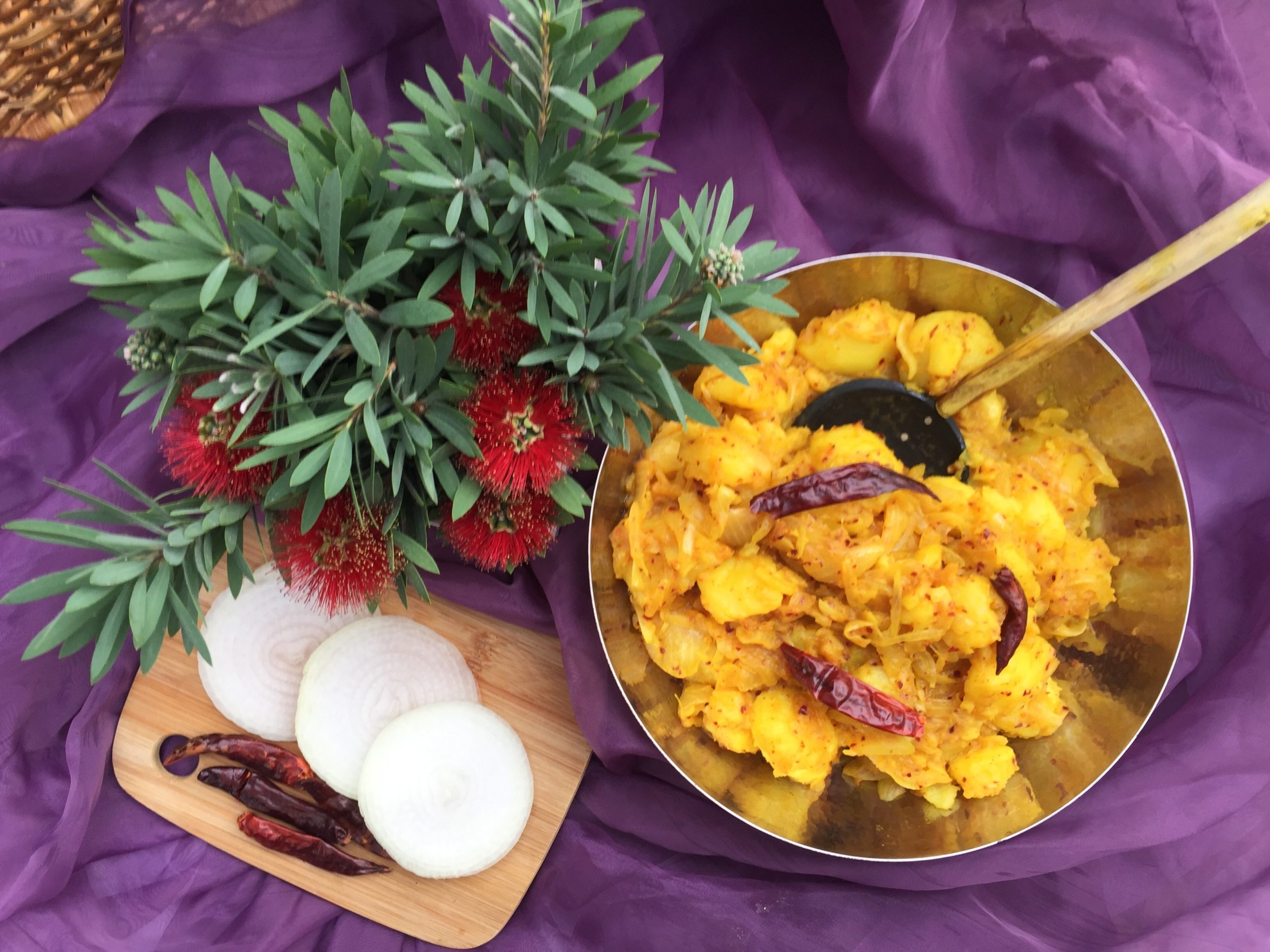 A bowl of curried potatoes, a small cutting board with onion slices and peppers, and a green and red plant.