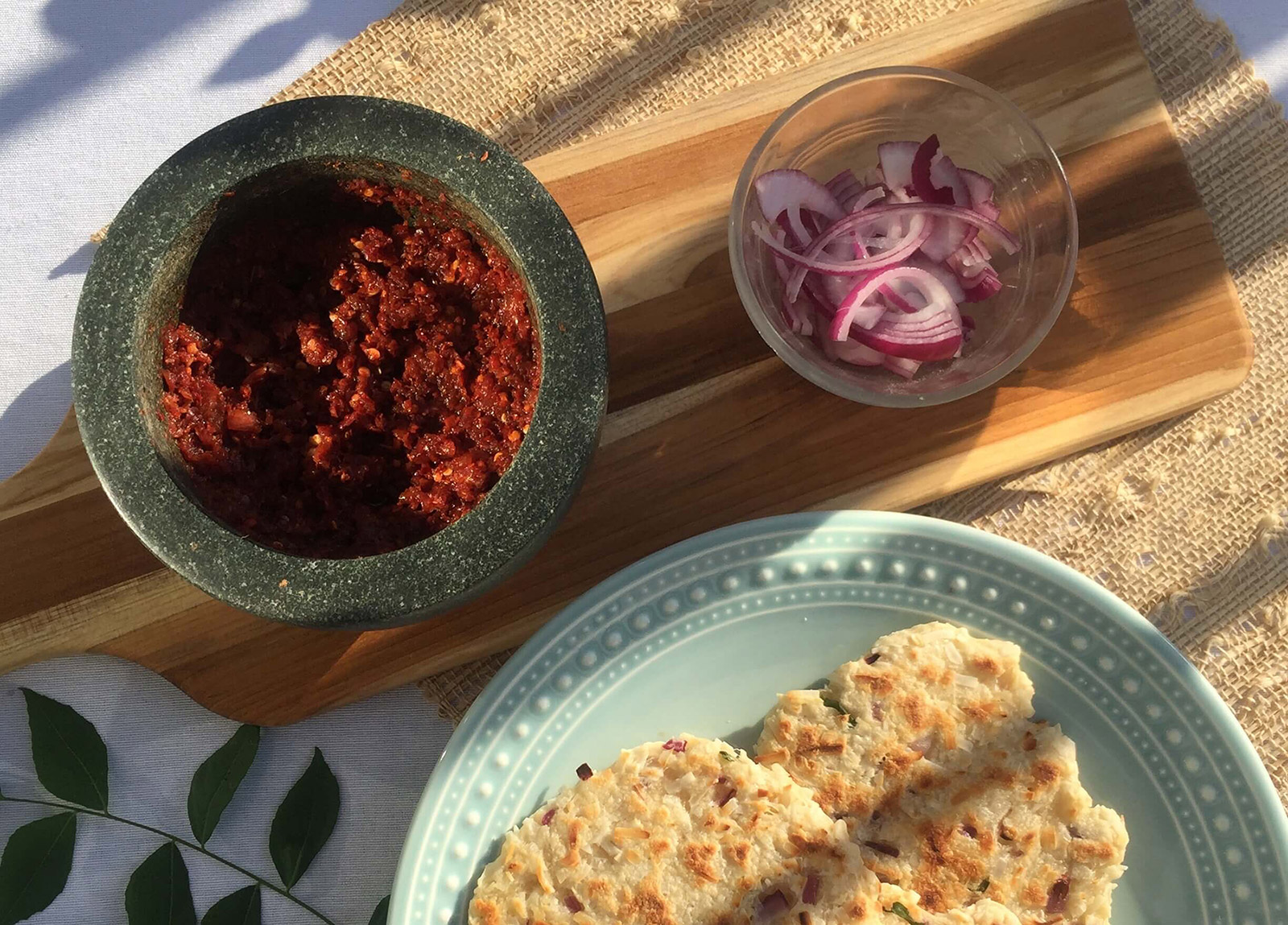 Roti served on a plate. Mortar containing chili and onion sambol. Small dish of sliced onion and sprig of curry leaf as decorative elements.